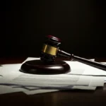 A wooden gavel resting on a stack of legal documents on a desk under dramatic, low-key lighting.