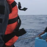 A Mexican navy sailor in an orange life vest hauls a rope toward two fishermen sitting inside a pale blue fishing cooler on open Pacific swells