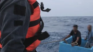 A Mexican navy sailor in an orange life vest hauls a rope toward two fishermen sitting inside a pale blue fishing cooler on open Pacific swells