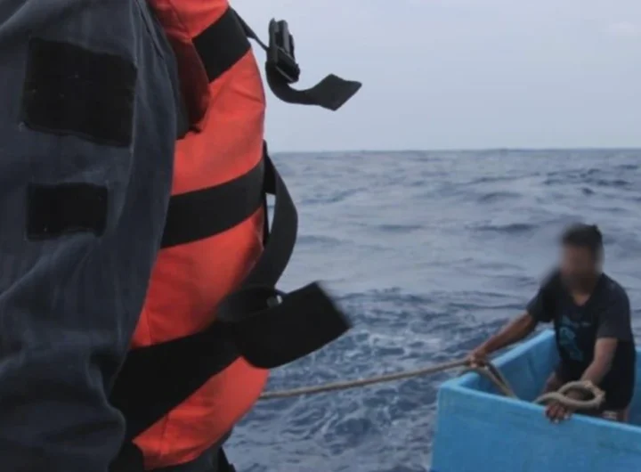 A Mexican navy sailor in an orange life vest hauls a rope toward two fishermen sitting inside a pale blue fishing cooler on open Pacific swells